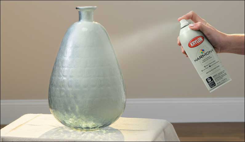 A person spraying a vase with a can of Krylon Harmony spray paint.